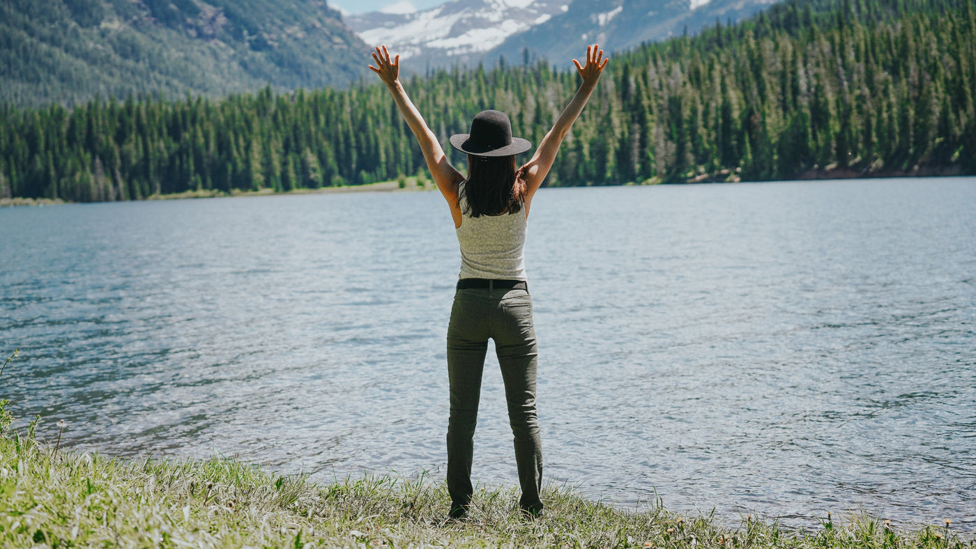 Woman with her hands in the air looking upon a lake. The woman is wearing a JeltX Adjustable belt with hiking pants, tank top and hat.